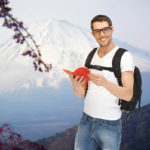 people, travel, tourism and education concept - happy young man in eyeglasses with backpack and book travelling over japan mountains background